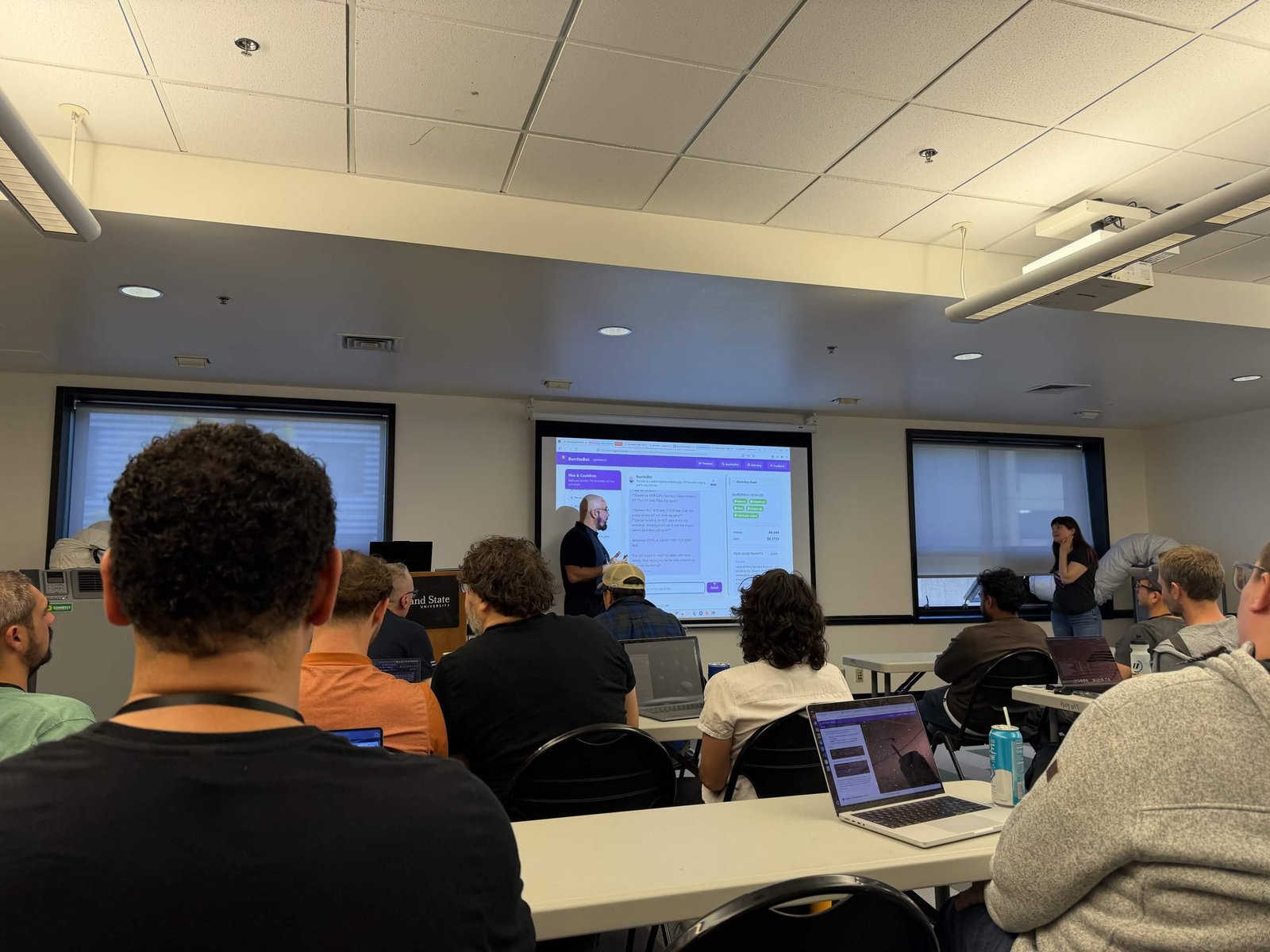 A workshop room at DevOpsDays Portland 2026, attendees on laptops, Michael at the screen running the Unleash an Agent exercise with Whitney Lee at right